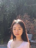 A young asian woman with long reddish-brown hair wears a pink t-shirt and gives a small smile to the camera in front of a large plant and bench in the background.